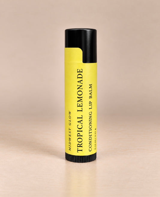 A single tube of Midwest Glow Tropical Lemonade Conditioning Lip Balm, featuring a bright yellow label and black cap, standing upright on a light beige surface against a matching beige background.